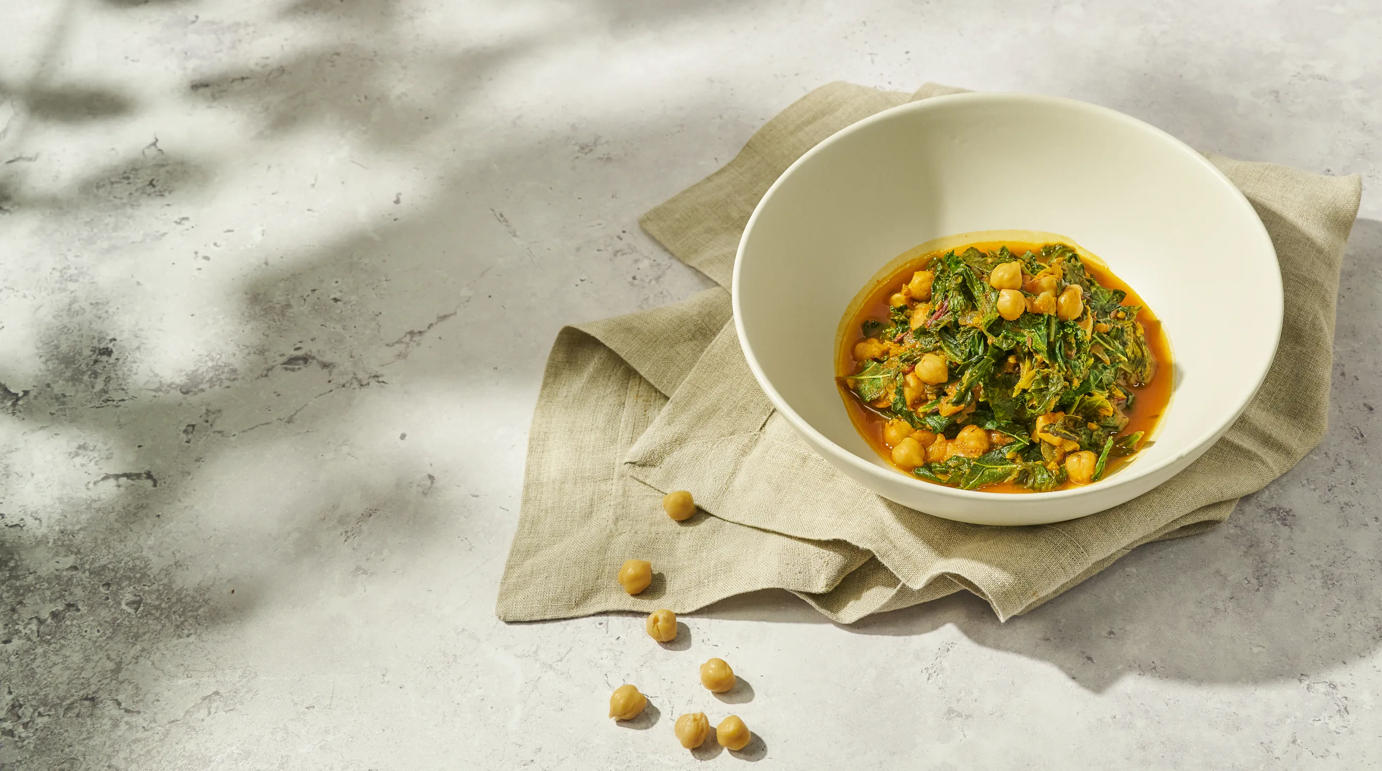 A bowl of chickpeas and braised greens in a warm turmeric broth, resting on linen in soft dappled light